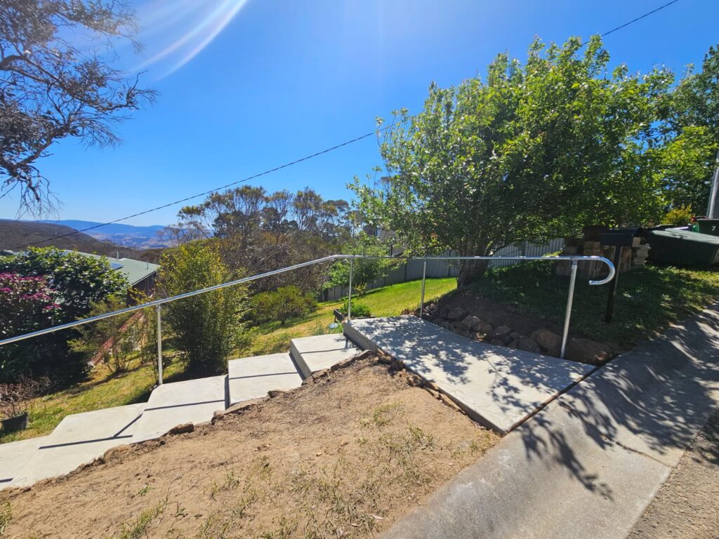 A series of concrete outdoor steps with Uni-fit silver metal handrails descends a grassy slope