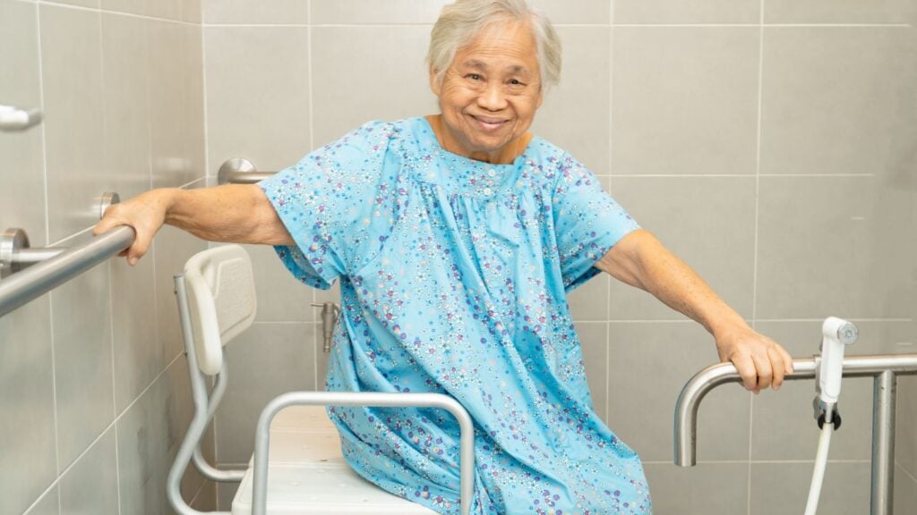 Elderly woman in a hospital gown holding stainless steel grab rails on both sides in a tiled bathroom