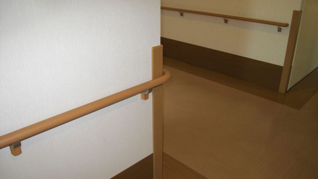 Timber wall-mounted handrail with wall brackets running along a corridor in a medical facility