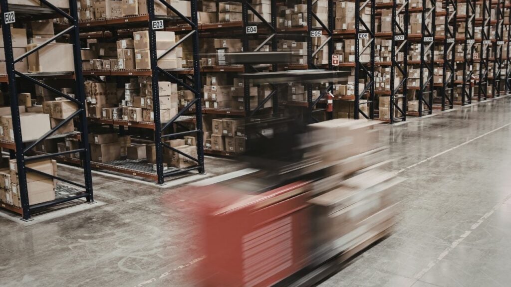 A forklift moving through a warehouse aisle lined with steel shelving and stacked cardboard boxes